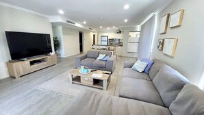 Una sala de estar moderna con un televisor de pantalla plana, sofás grises, una mesa de centro y una cocina al fondo.