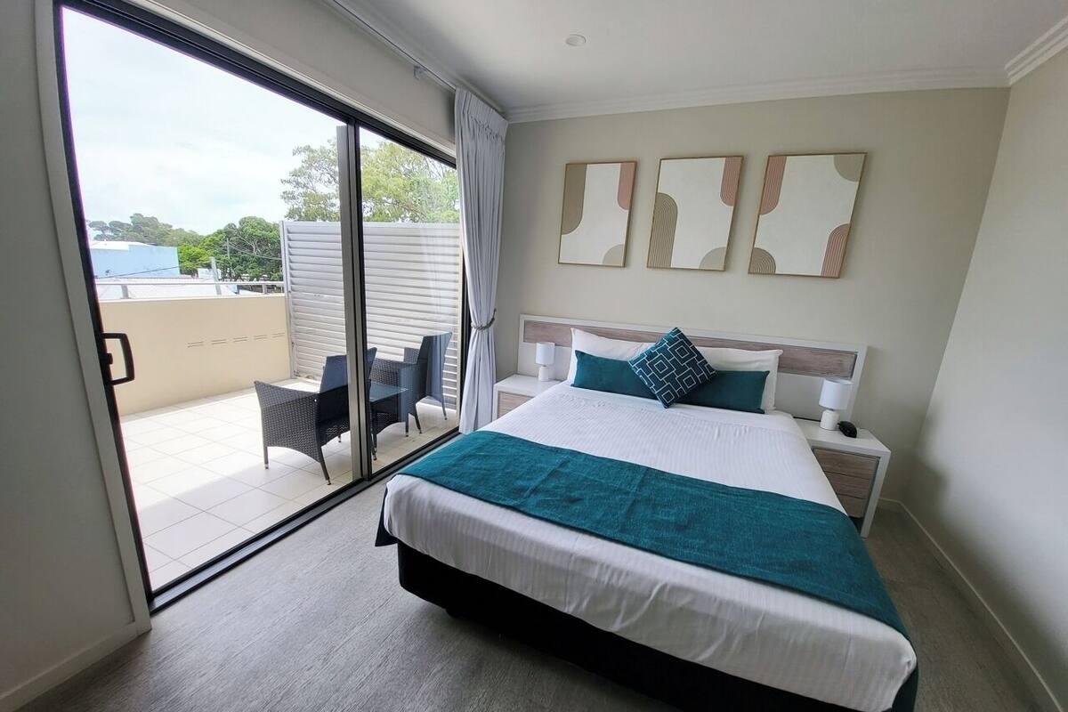 A modern hotel room with a large bed, a sliding glass door to a balcony, and a view of the outside.