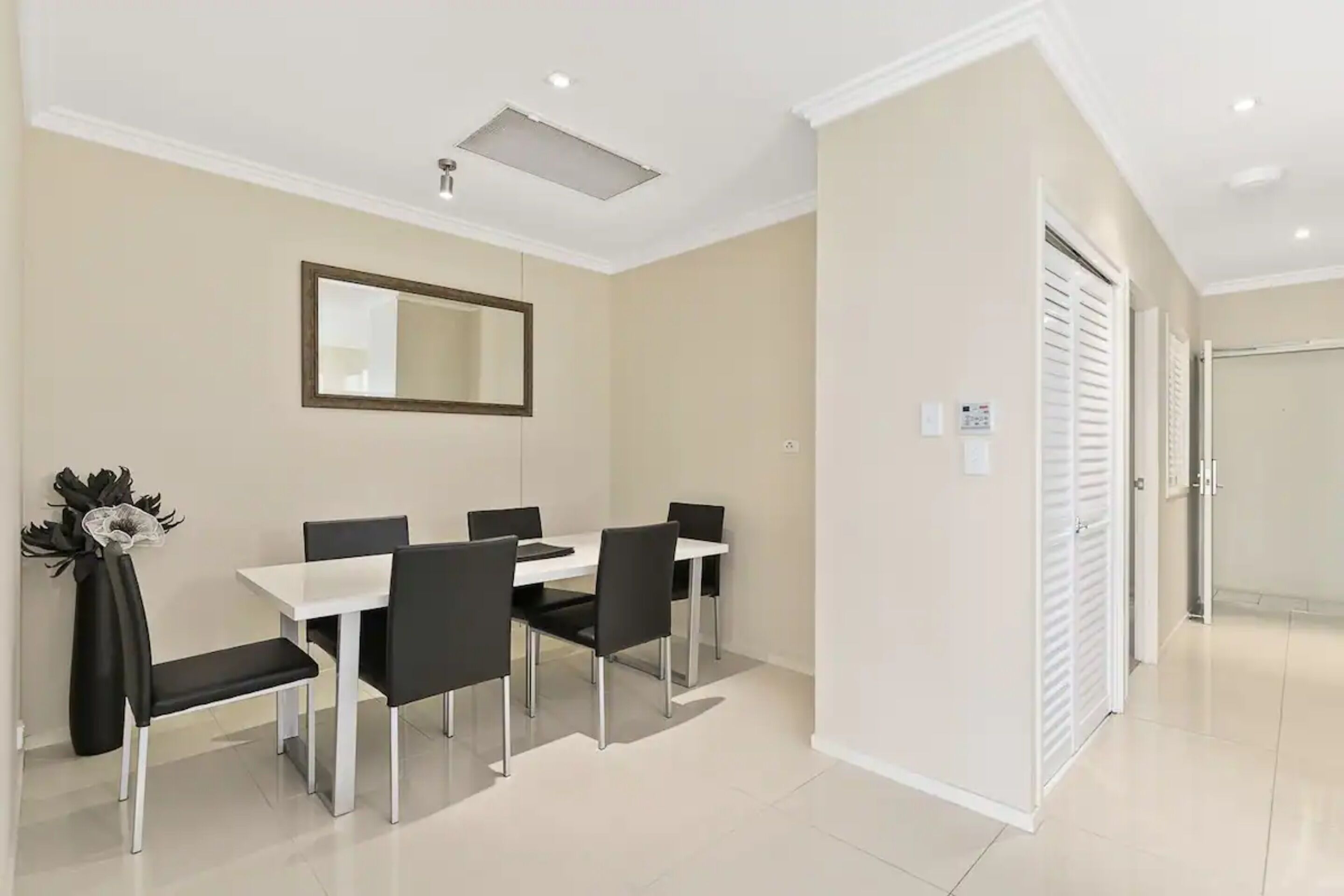 A modern dining area with a white table and black chairs, a large mirror on the wall, and a vase with flowers.