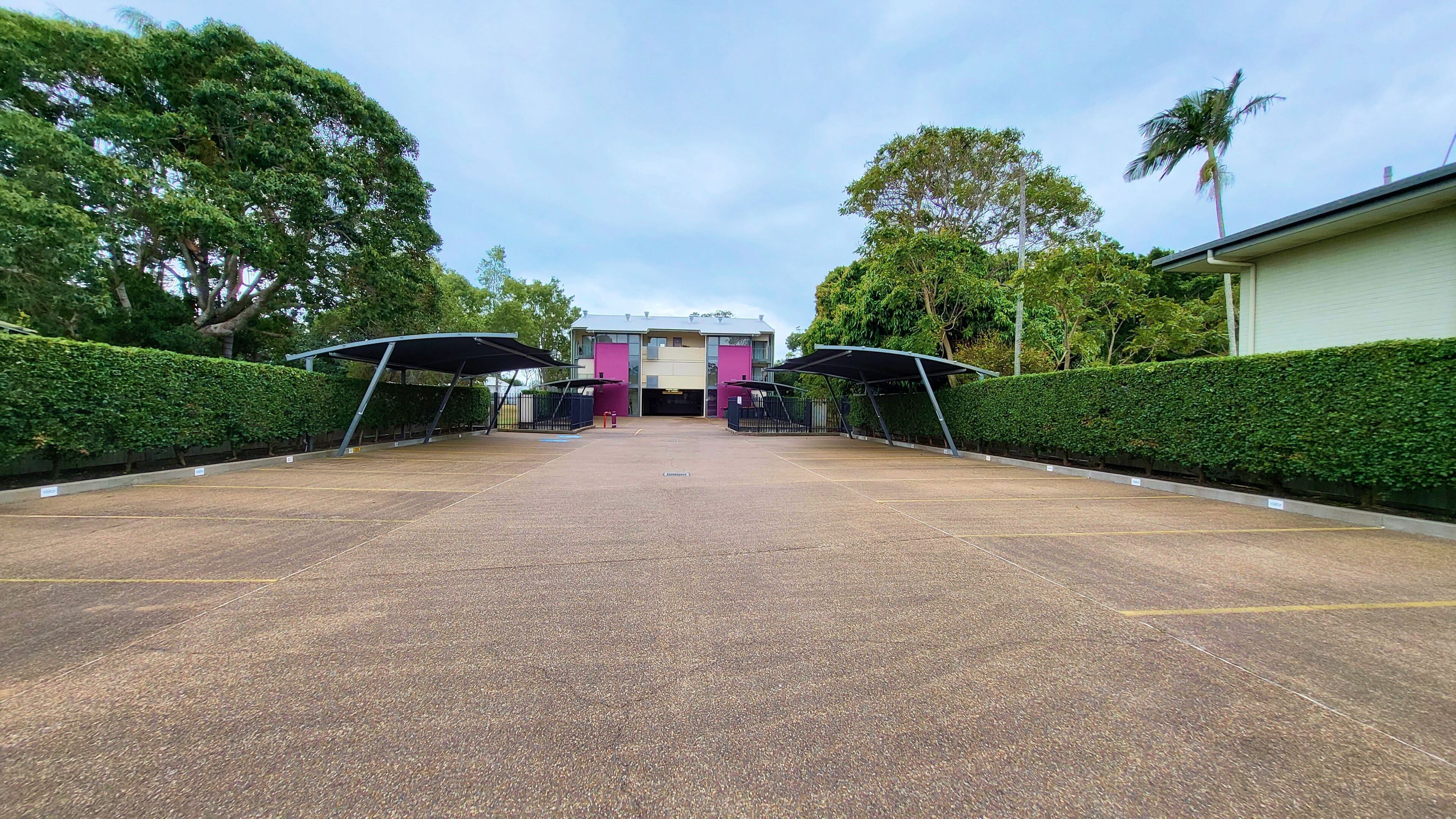 A parking area with a covered walkway leading to a building with a modern design, surrounded by greenery.