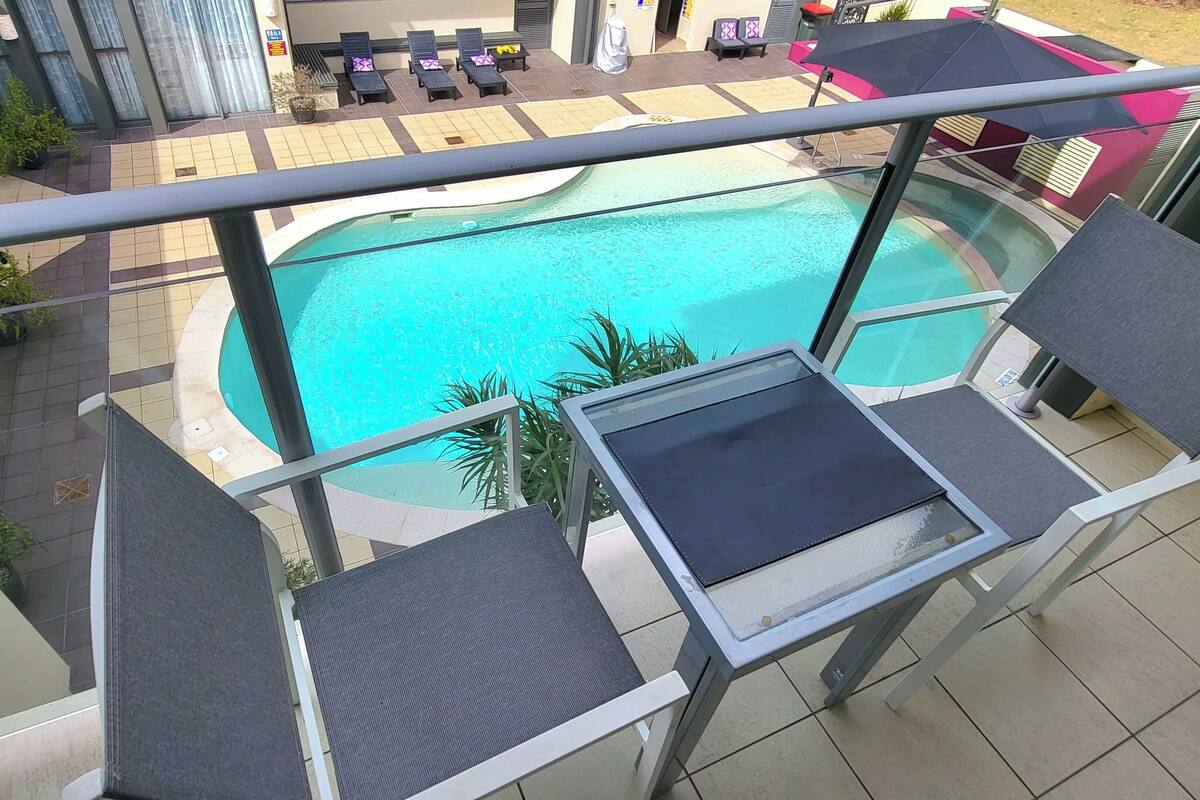 A balcony with a view of a swimming pool, lounge chairs, and a sun umbrella.