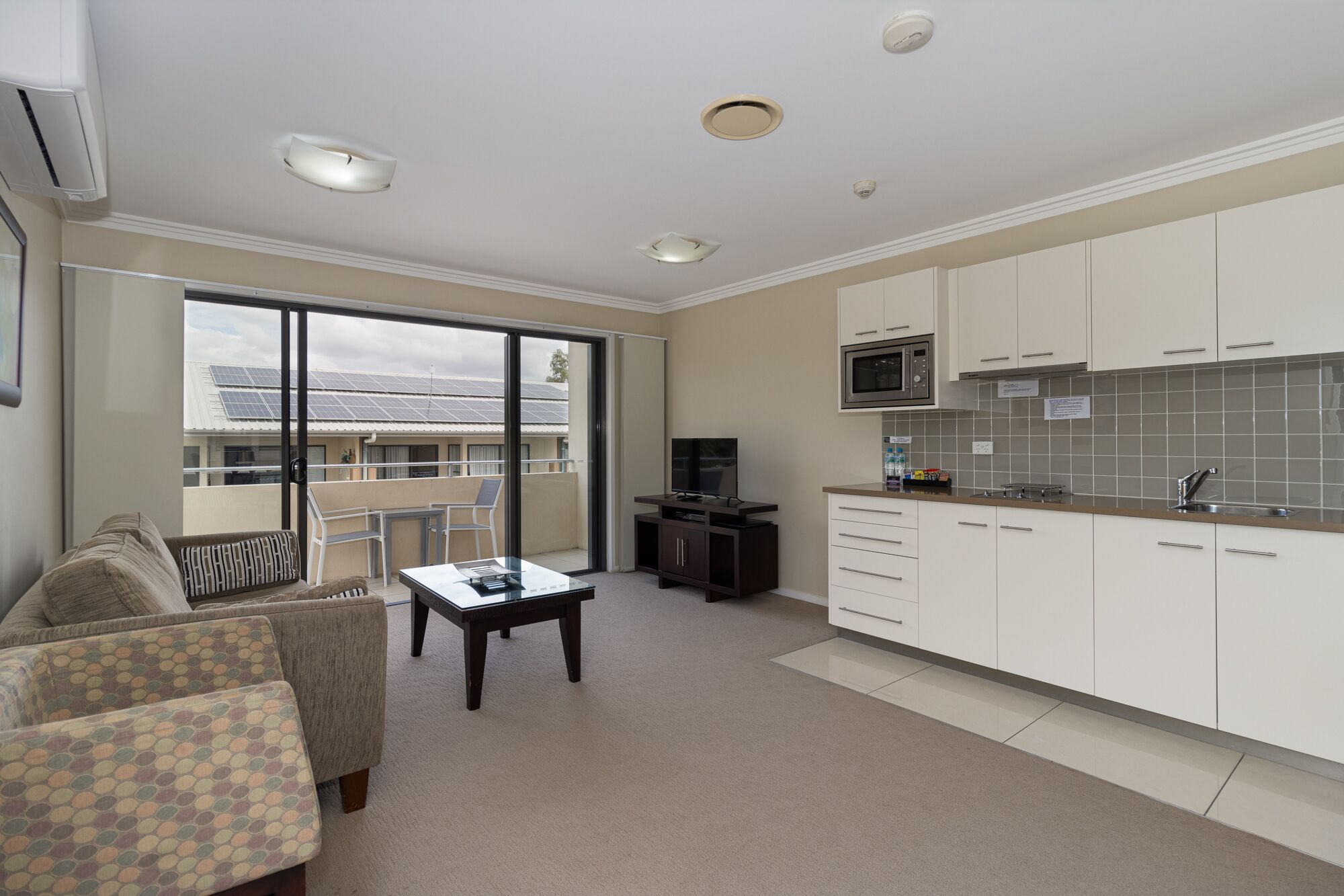 Un apartamento moderno con balcón, cocina con armarios blancos, comedor con mesa y sillas, y televisor.