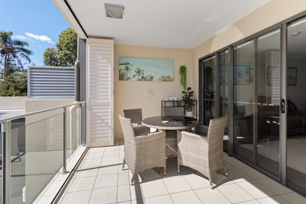 A balcony with a round table and wicker chairs, a sliding glass door, and a painting of a tropical scene.