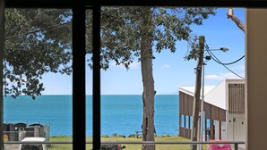 Vista a través de una ventana de un edificio con vista a una playa, cielo despejado, algunos árboles y una masa de agua.