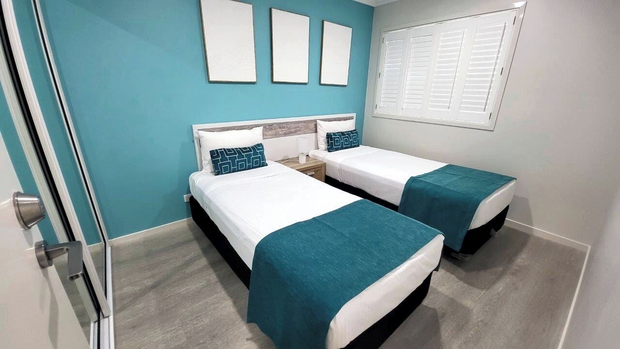 A hotel room with two beds, teal bedding, a wooden headboard, a window with blinds, and a teal-colored carpet.