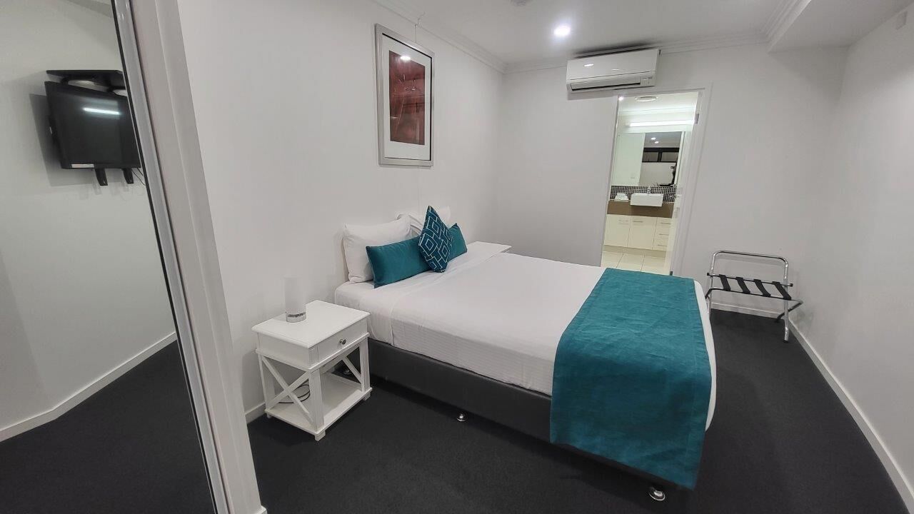 Habitación de hotel con cama, mesita de noche, aire acondicionado de pared y puerta que da a un baño.