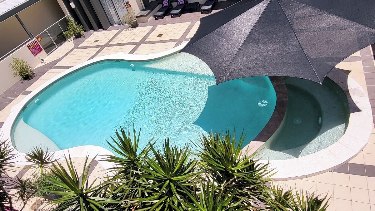 Una piscina de forma singular, rodeada por una terraza con baldosas y protegida por un gran paraguas.