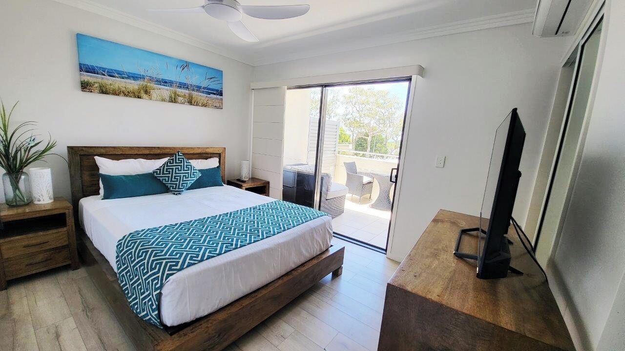 A bedroom with a bed, a nightstand, a TV, a ceiling fan, and a balcony with outdoor furniture.