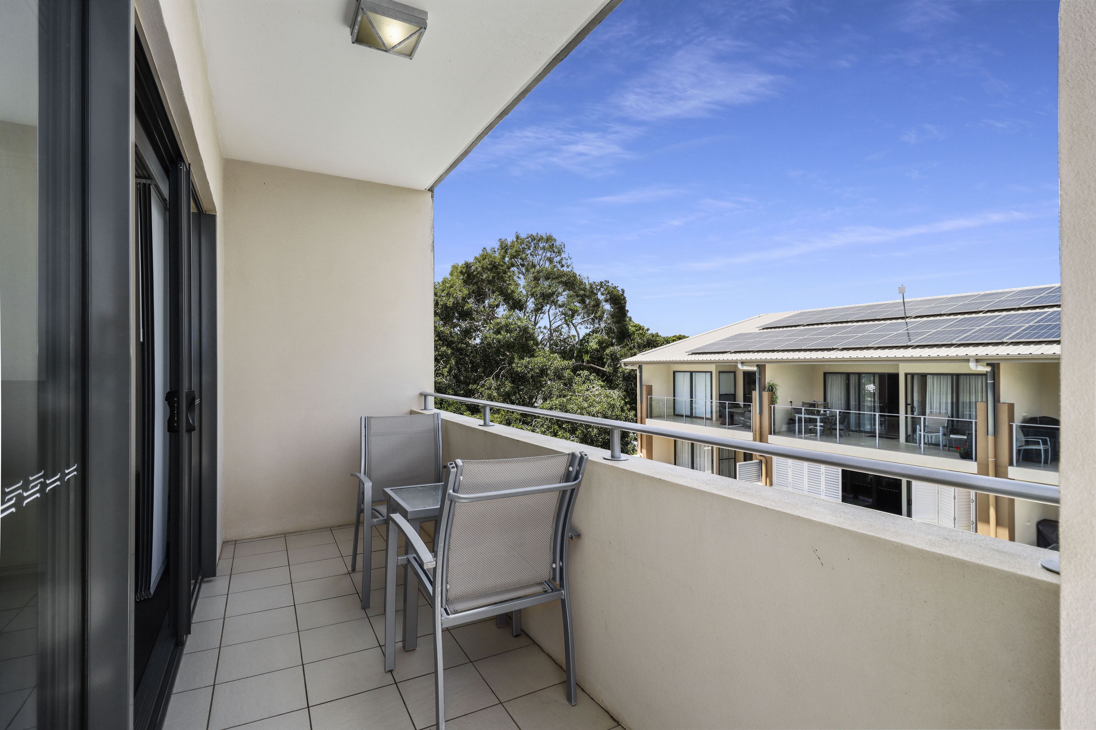 A balcony with a table and chairs, a door, and a view of a building with solar panels.