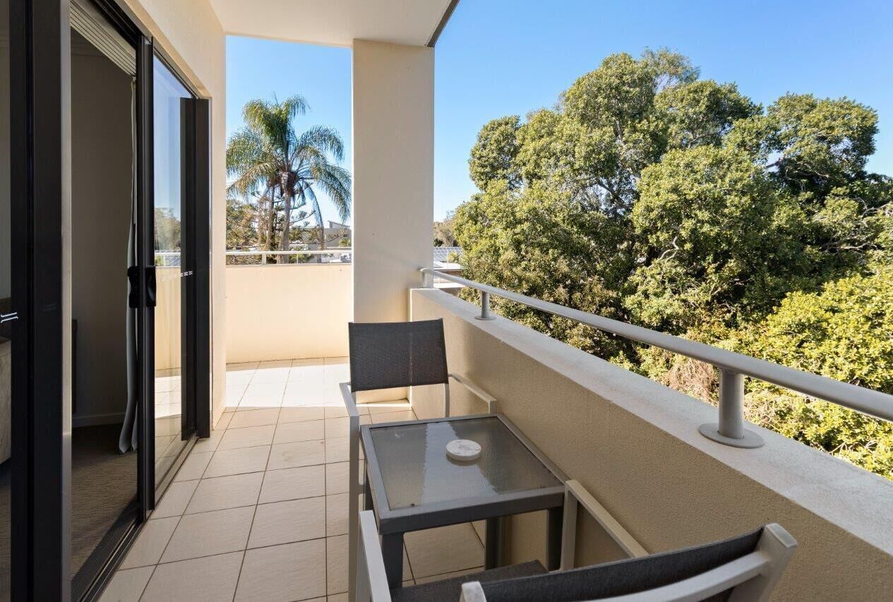 A balcony with a glass table and chairs, overlooking a lush green landscape with trees and a clear sky.