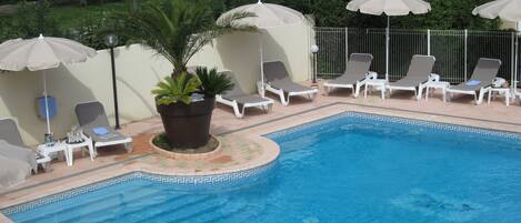 A swimming pool with lounge chairs and umbrellas, surrounded by greenery and a tiled border.