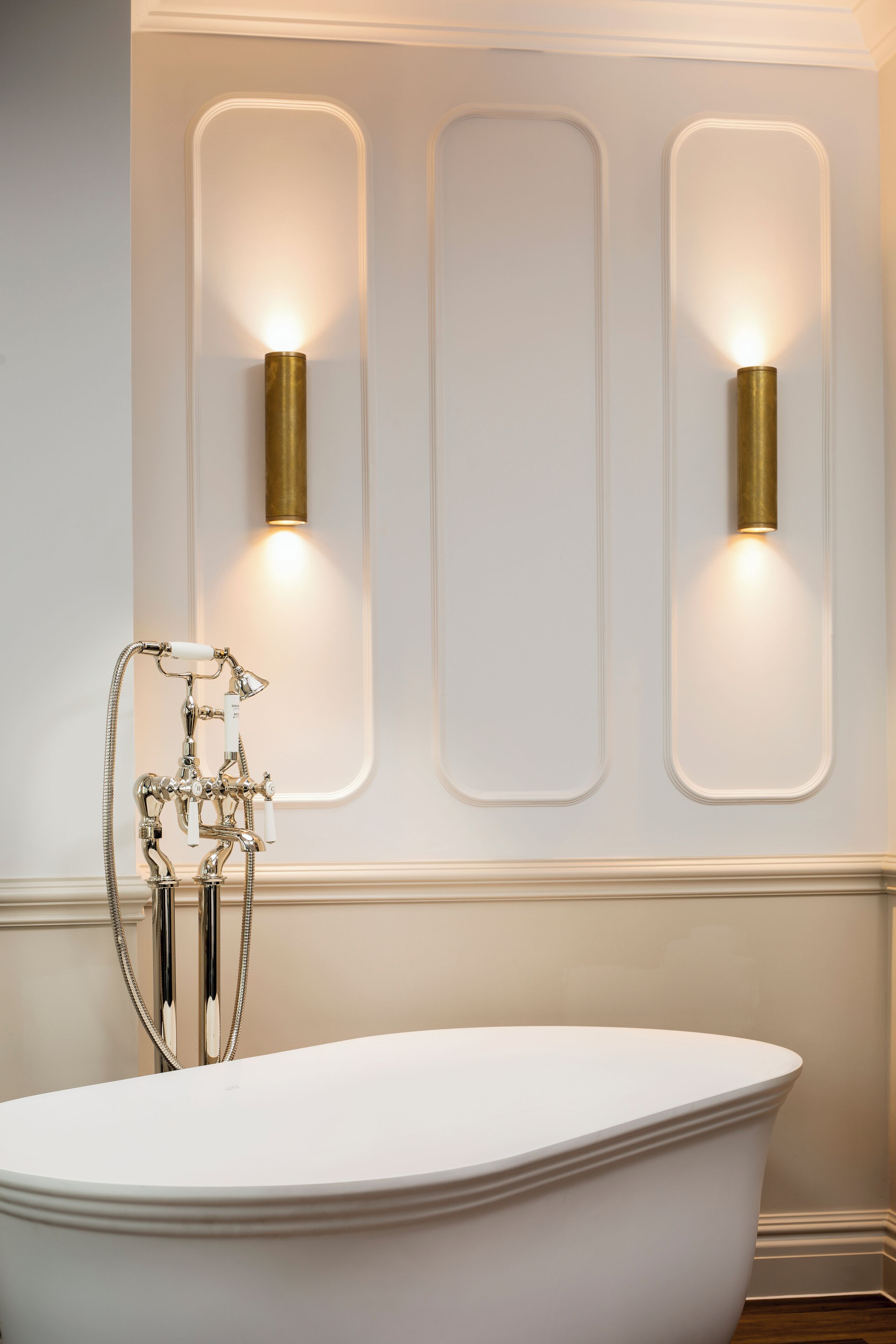 A white freestanding bathtub with a chrome faucet and hose, positioned in front of a wall with two wall-mounted sconces and a recessed panel.