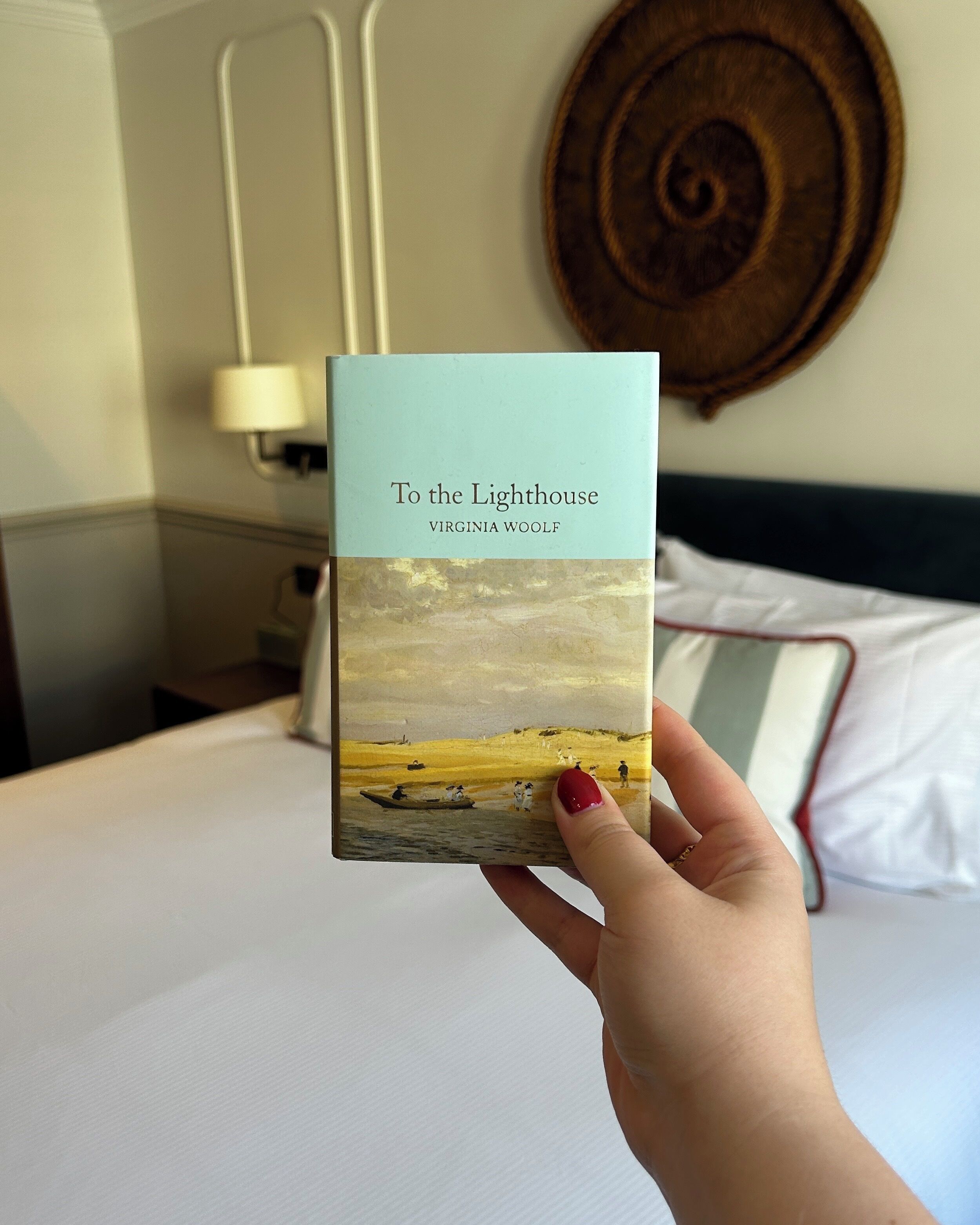 A hand holding a book titled "To the Lighthouse" by Virginia Woolf, with a painting of a coastal scene on the cover.