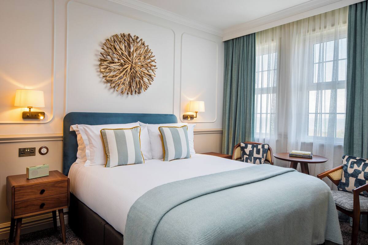 A hotel room with a large bed, two bedside lamps, a decorative wall piece, a window with curtains, and a small table with chairs.