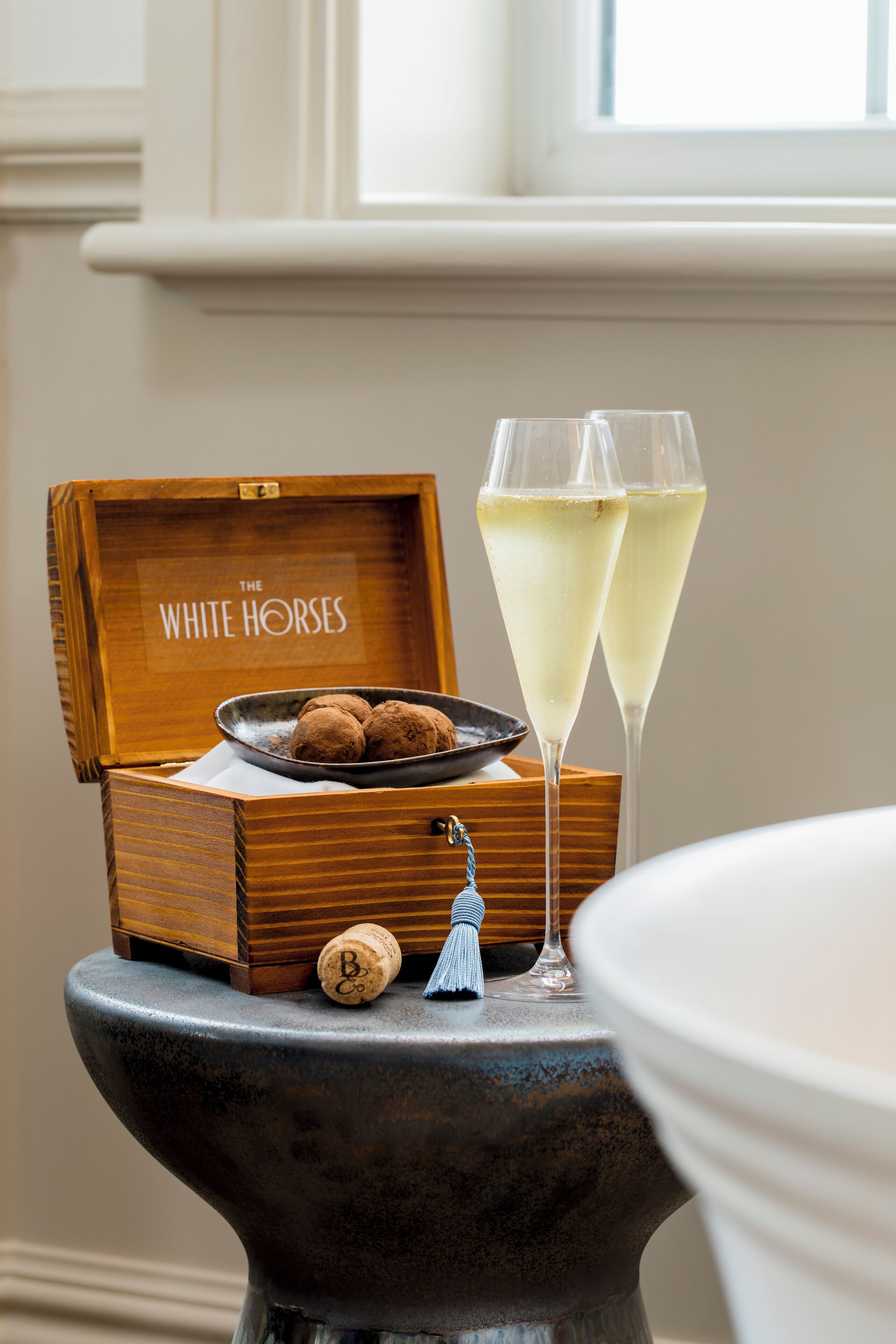 A wooden box with a label "The White Horses" on a table with two champagne glasses and a bowl of chocolates.