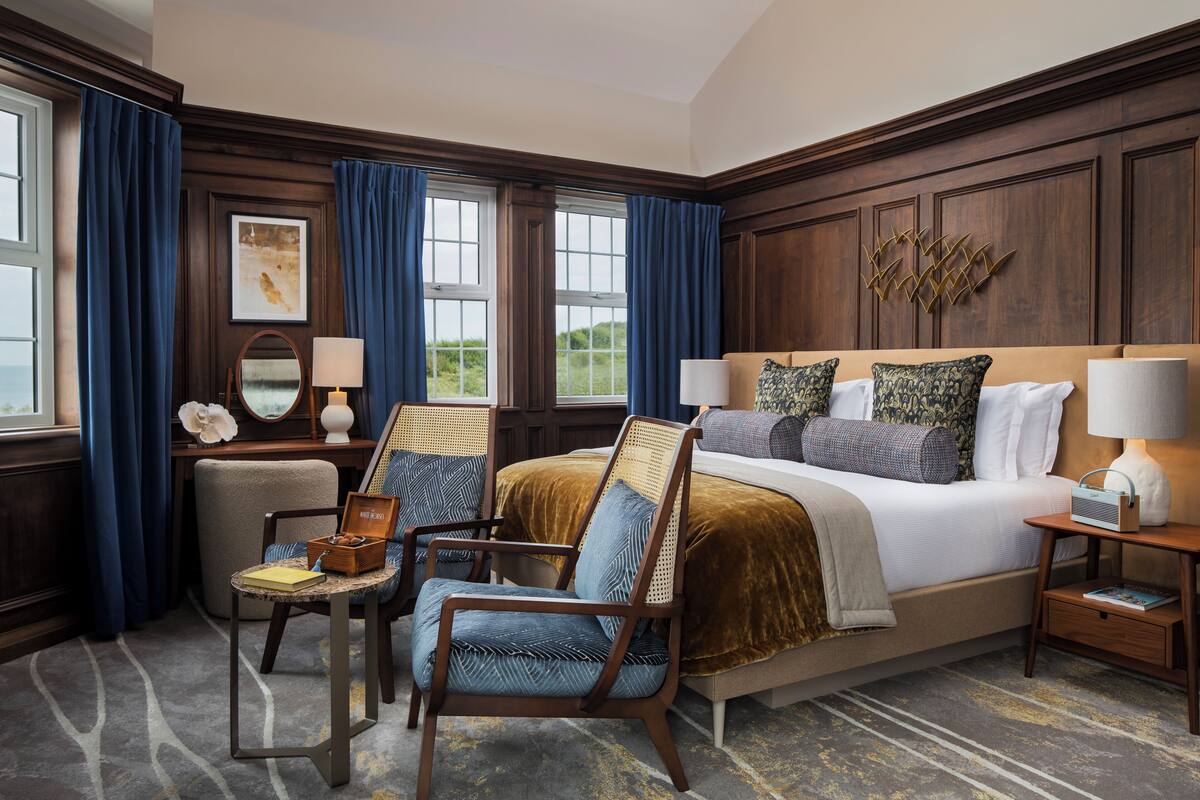 A hotel room with a large bed, two armchairs, a nightstand, a lamp, a mirror, and a view of the outdoors.