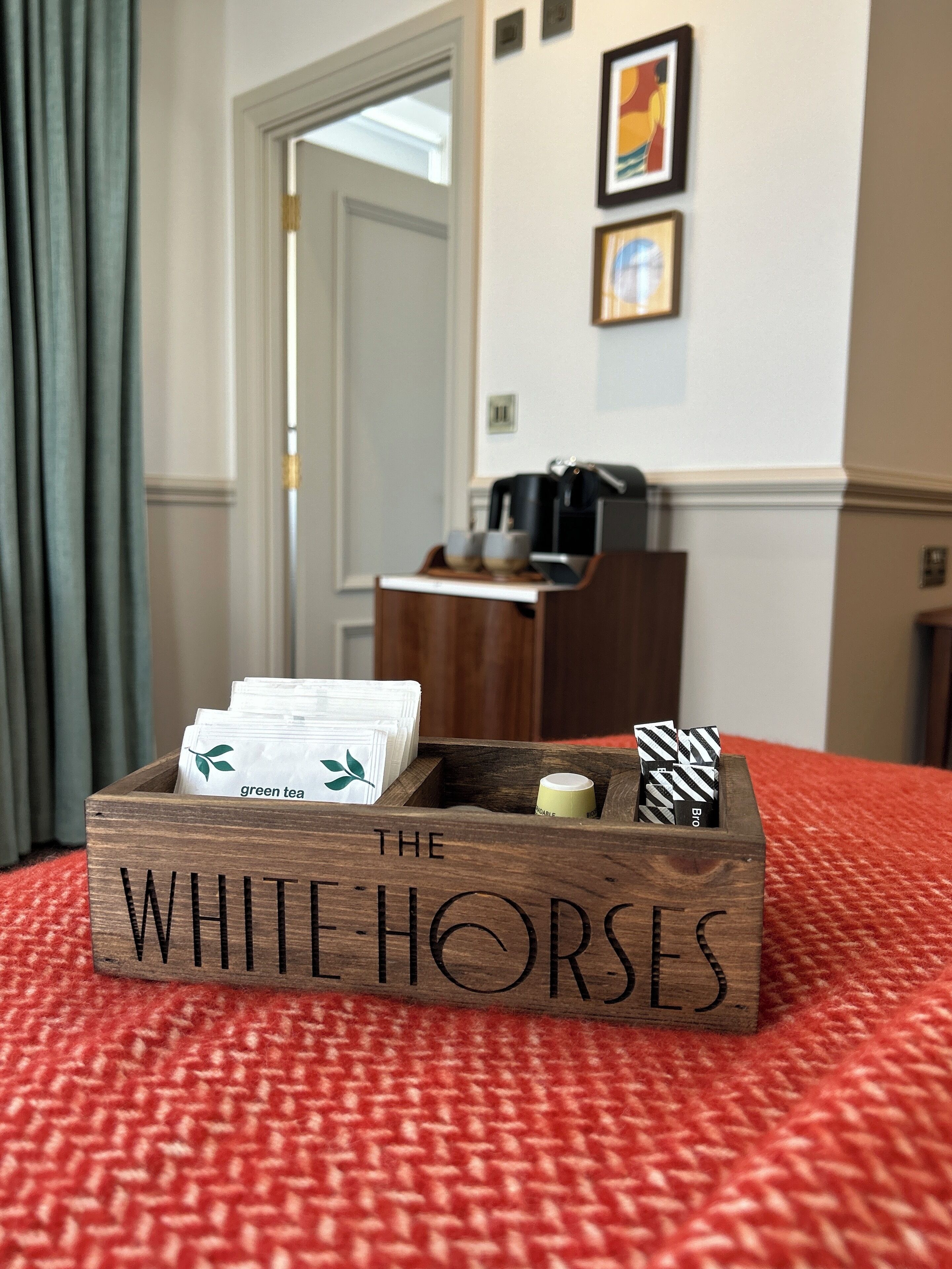 A wooden box labeled "The White Horses" on a red carpet, containing tea bags and a small container.