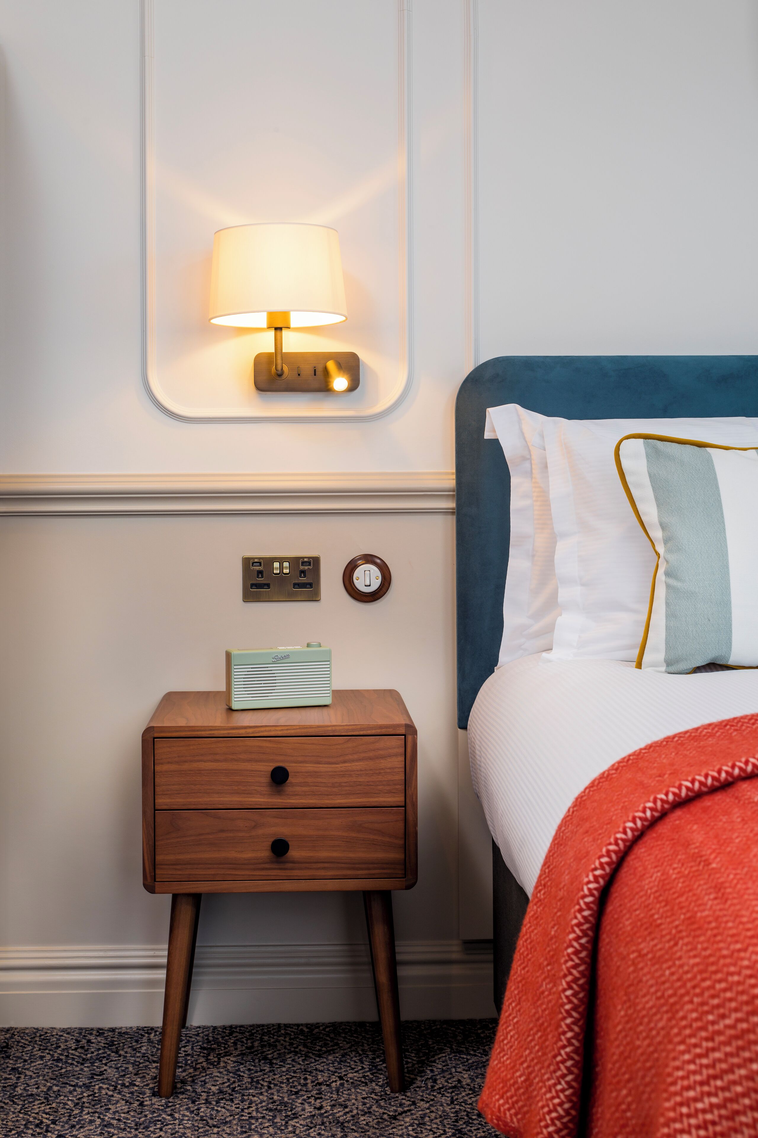 A neatly arranged hotel room with a bedside table, a lamp, a radio, and a bed with a blue headboard and white bedding.
