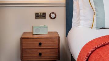 A neatly arranged hotel room with a bedside table, a lamp, a radio, and a bed with a blue headboard and white bedding.