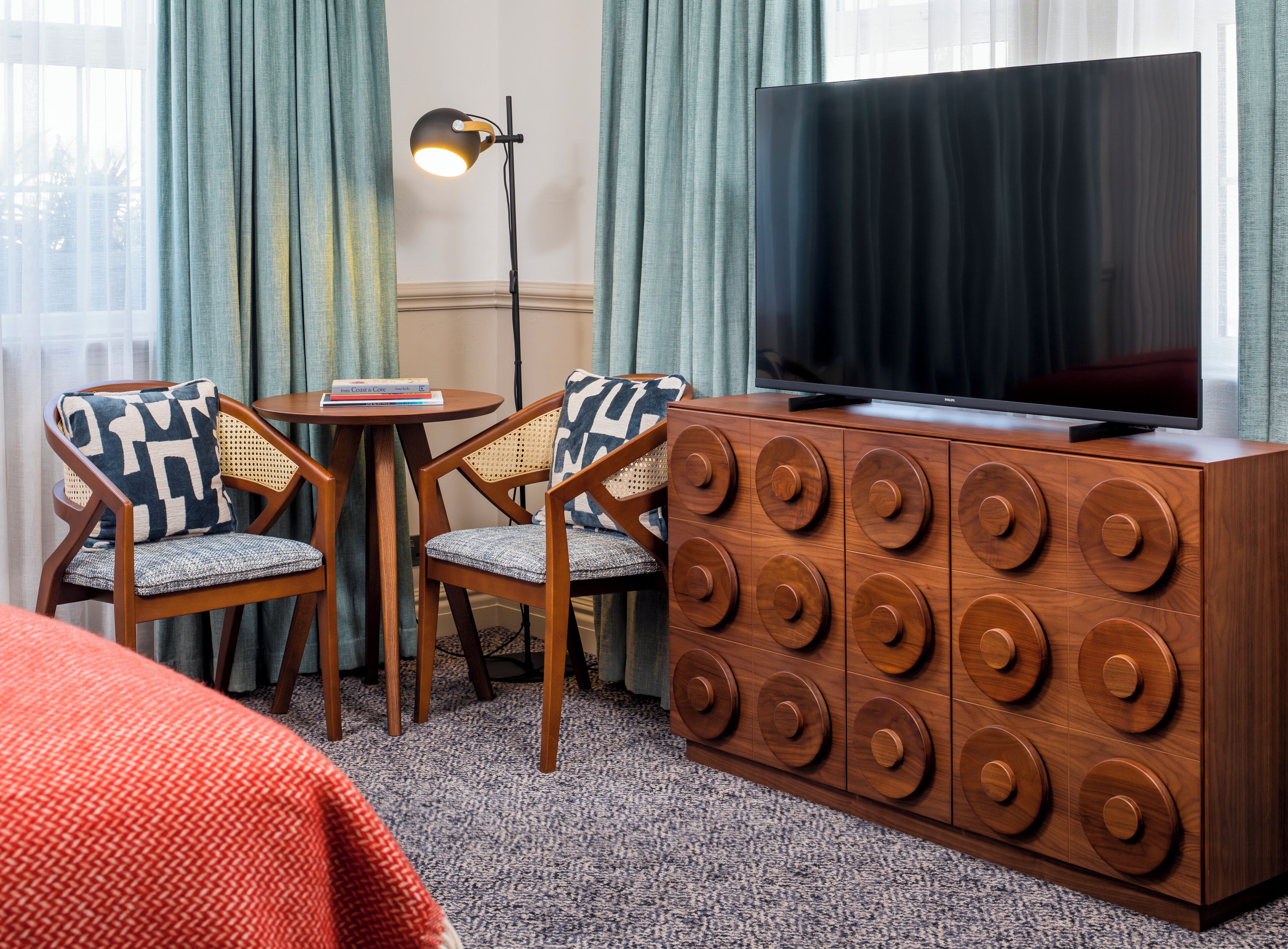 A hotel room with a large flat-screen TV, two wooden chairs with patterned cushions, a small round table, and a wooden cabinet with circular handles.