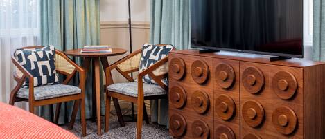 A hotel room with a large flat-screen TV, two wooden chairs with patterned cushions, a small round table, and a wooden cabinet with circular handles.