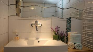 Un baño con un lavamanos blanco, un espejo de marco ondulado, un jarrón verde con flores y un perchero con toallas dobladas.