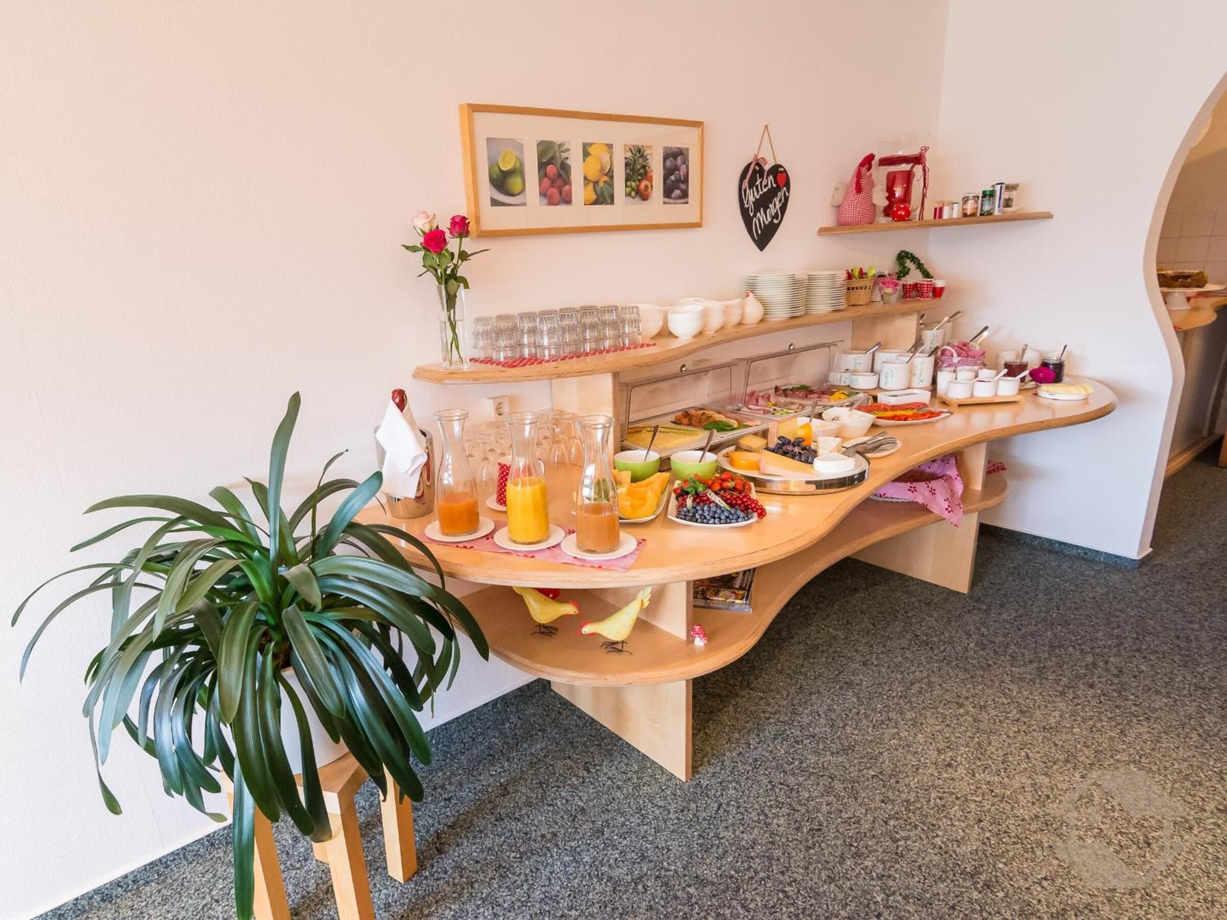 A buffet setup with various food items and drinks, a potted plant, and a framed picture on the wall.