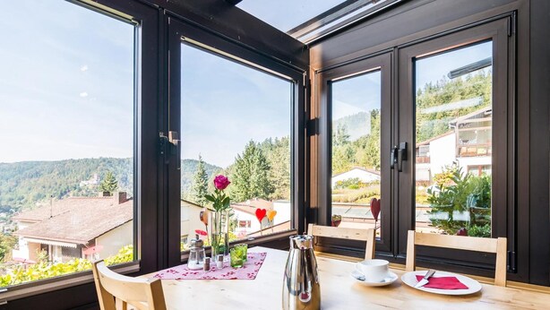 A dining area with a table set for two, large windows offering a view of a mountainous landscape. - Aparthotel Hochwald (Bad Liebenzell)
