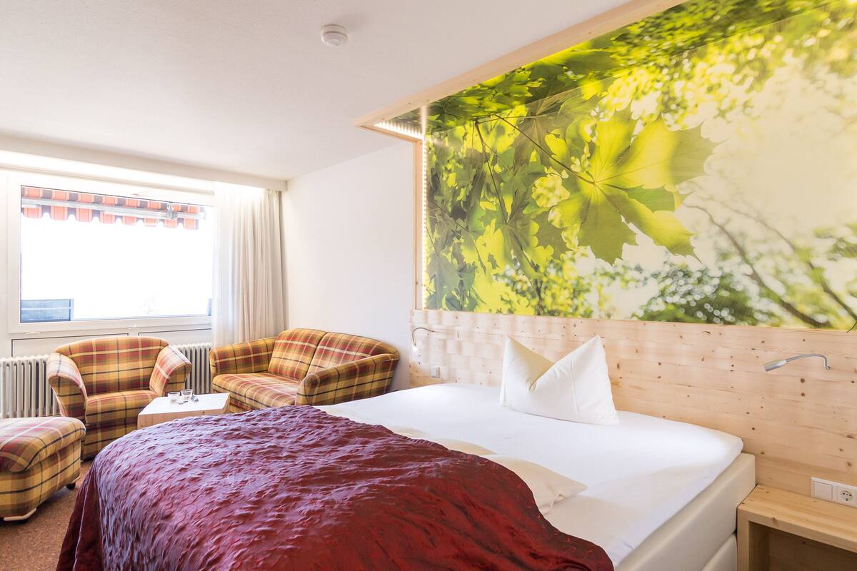 A hotel room with a bed, two plaid armchairs, a small table, and a large window.