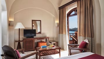 A hotel room with a bed, a desk with a television, a chair, a table with fruit, and a view of the sea.