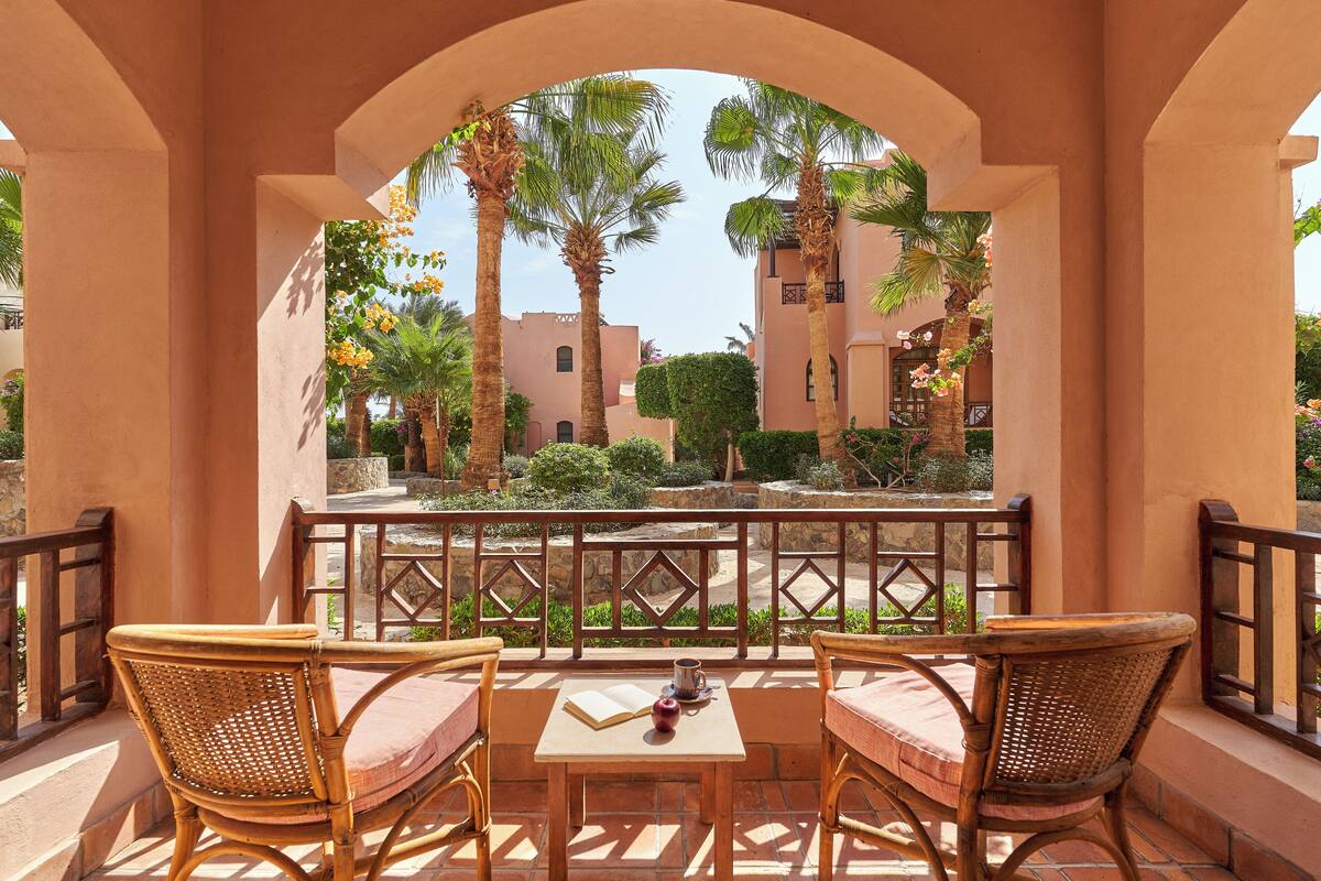 A balcony with two wicker chairs and a table, overlooking a courtyard with palm trees and buildings.