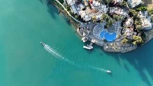Vista aérea de un complejo turístico frente al mar, con un puerto deportivo, piscina y varios edificios con balcones.