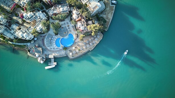 Vista aérea de un complejo turístico frente al mar, con piscina central, tumbonas y un muelle para embarcaciones.