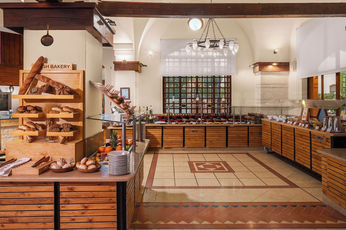 Een bakkerijbalie met verschillende broden en gebak, een zithoek met een patroon vloer, en uitzicht op het buiten via een raam.
