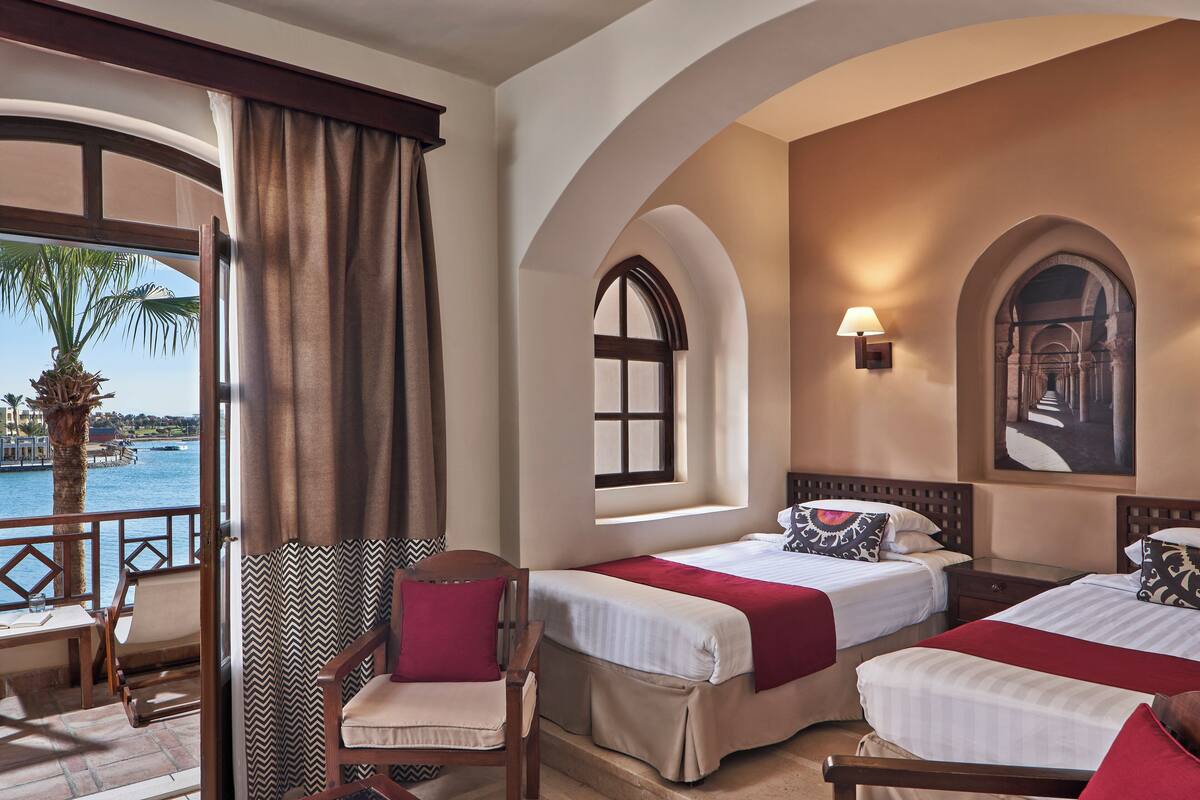 A hotel room with two beds, a chair, a table, and a view of the sea.