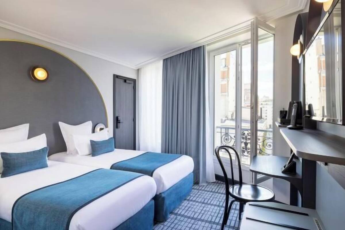 Ein Hotelzimmer mit zwei Betten, einem Schreibtisch, einem Sessel, einem großen Fenster mit Vorhängen und Blick auf die Stadt.
