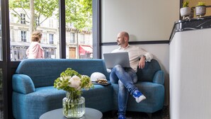 Un hombre sentado en un sofá azul, con una computadora portátil, un jarrón de flores y una mesita con un florero de vidrio.