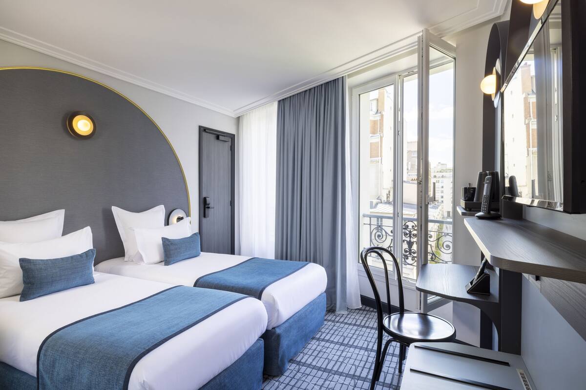Ein Hotelzimmer mit zwei Betten, einem Schreibtisch, einem Stuhl, einem Spiegel und Blick auf die Stadt durch das Fenster.