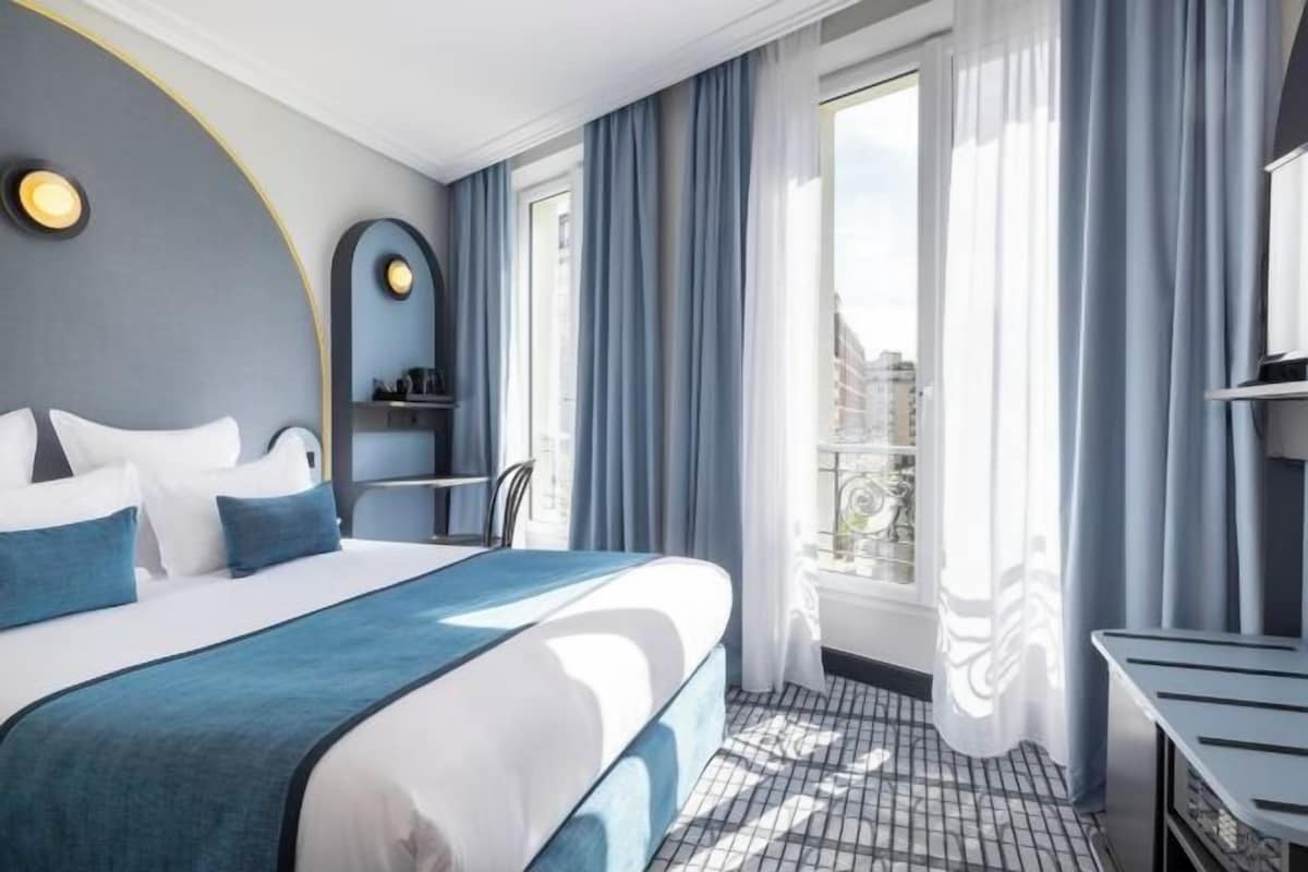 A hotel room with a bed, a desk, a chair, a window with curtains, and a view of buildings.