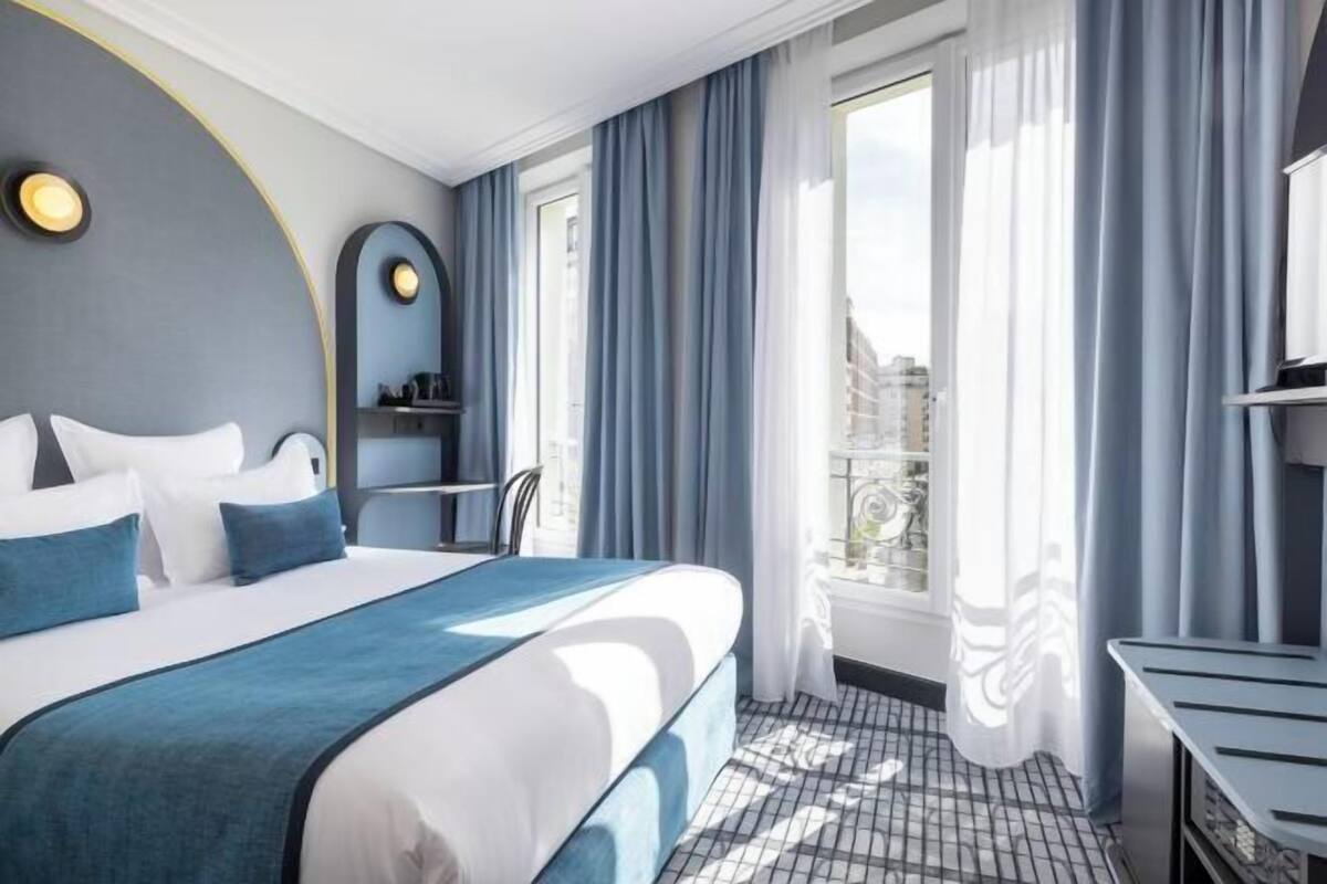 Ein Hotelzimmer mit Bett, Schreibtisch, Stuhl, einem Fenster mit Vorhängen und Blick auf Gebäude.