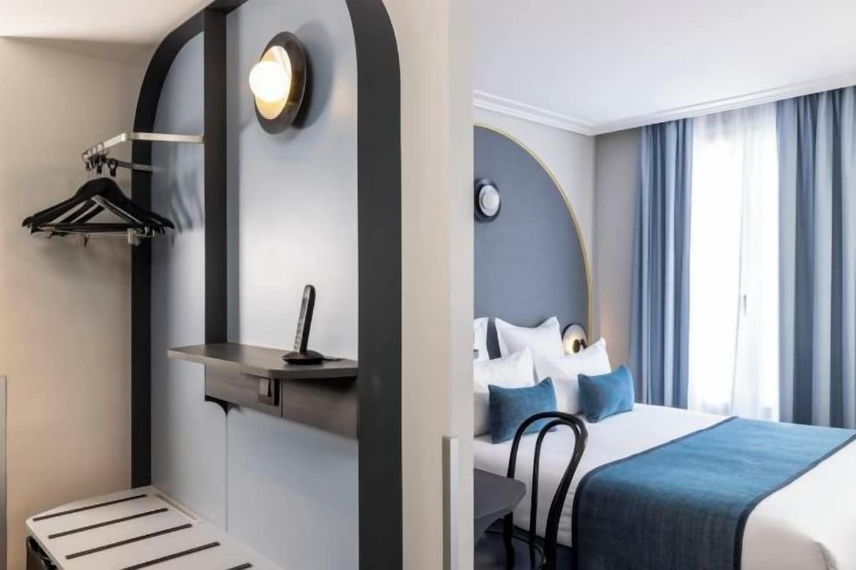 A hotel room with a bed, a desk, a chair, a hanging clothes rack, and a small table.