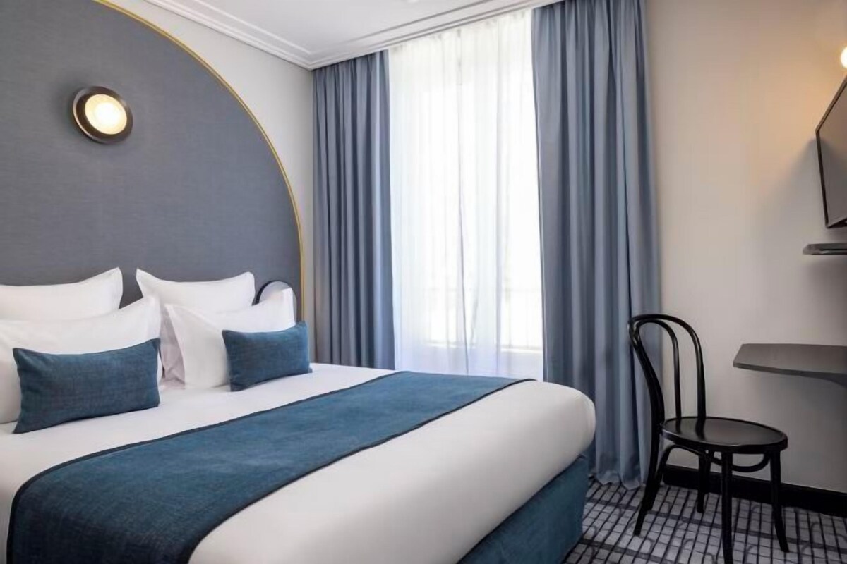 A hotel room with a large bed, a chair, a desk, and a window with curtains.
