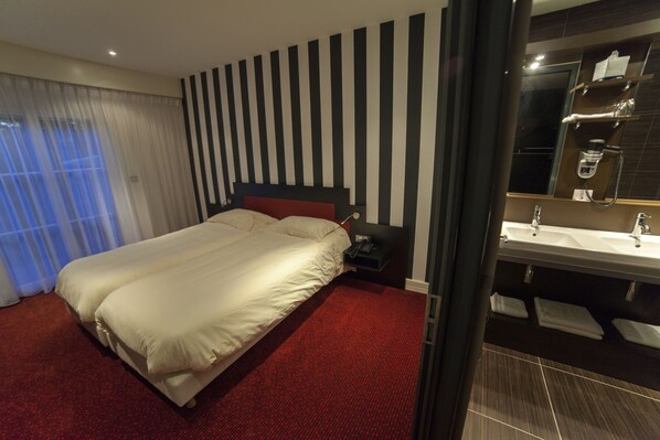 Habitación de hotel con una cama grande, alfombra roja, pared a rayas y baño con lavamanos y espejo.
