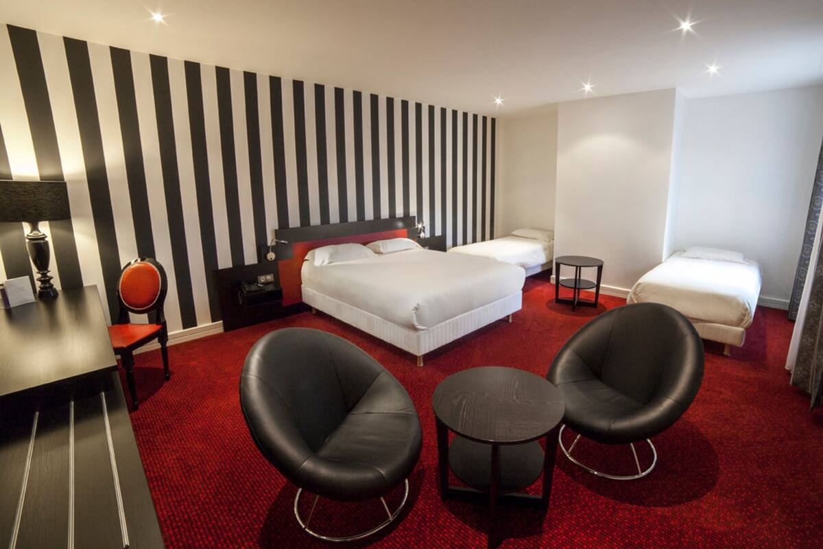 Ein Hotelzimmer mit einem großen Bett, zwei Einzelbetten, einem Schreibtisch mit Stuhl, zwei Sesseln, einem kleinen Tisch und einem an der Wand befestigten Fernseher.