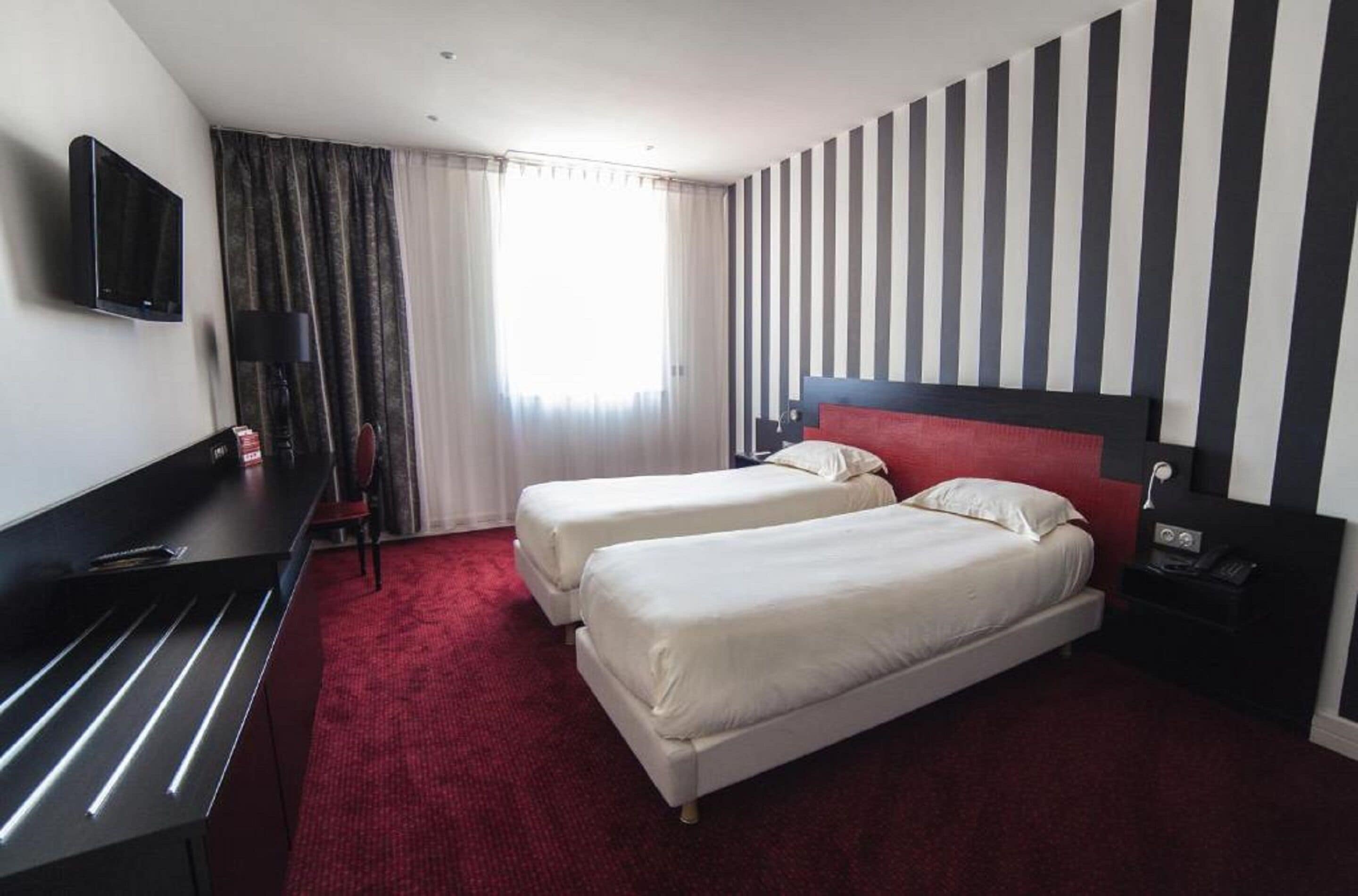 A hotel room with two beds, a desk, a TV, and a striped wall.