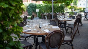 Een terras met ronde tafels en rieten stoelen, omringd door groen en gebouwen op de achtergrond.