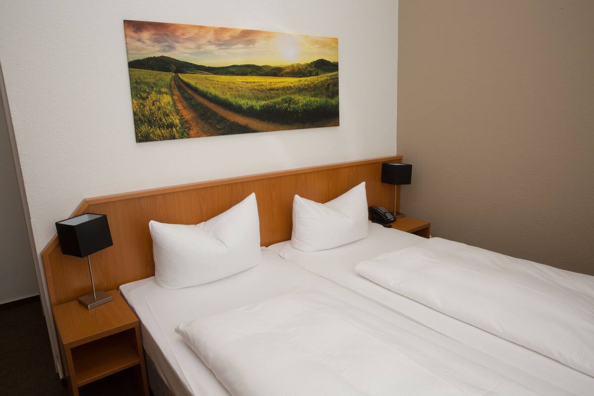A hotel room with a large bed, two bedside tables, a nightstand, a wall-mounted painting, and a wooden headboard.