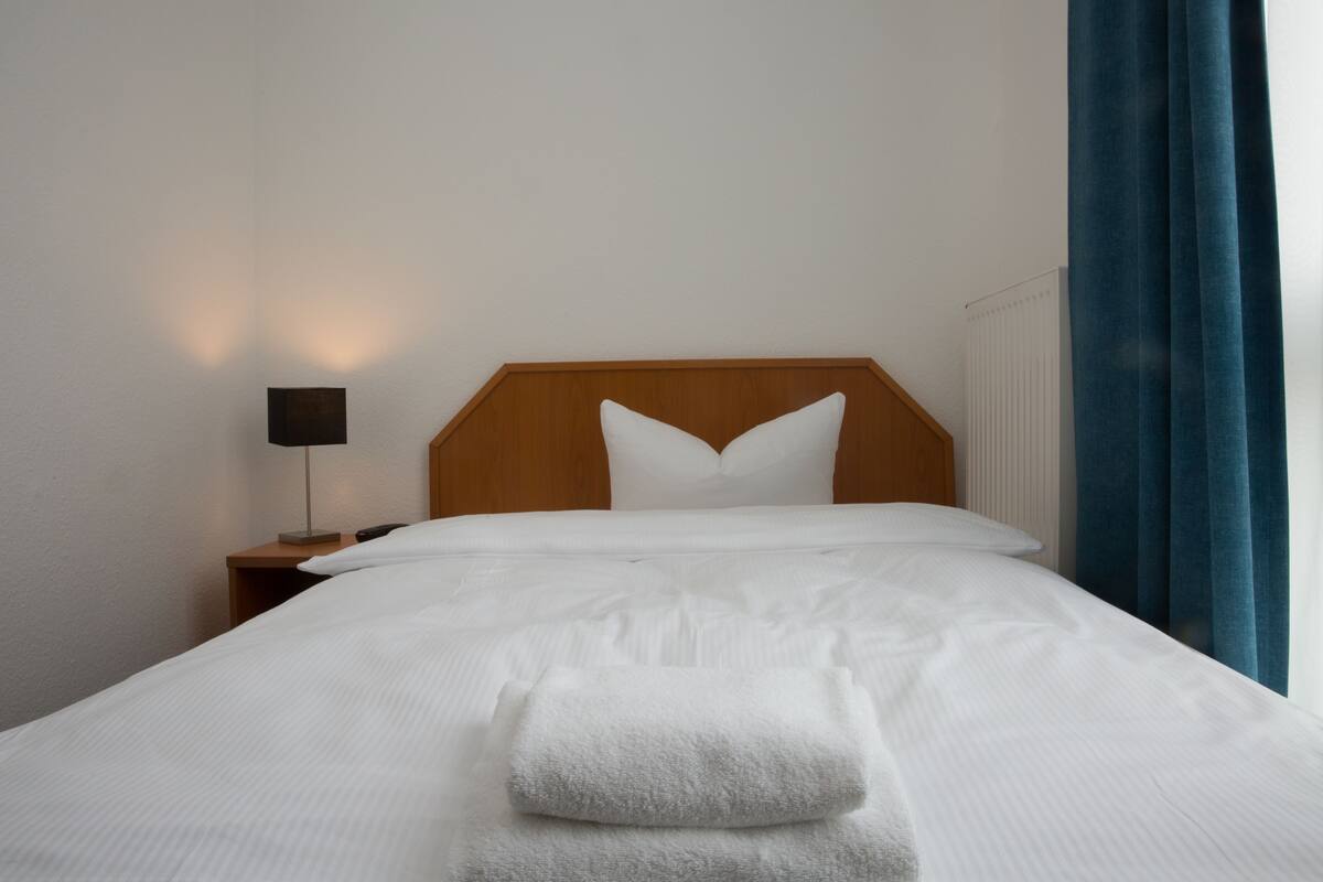 A neatly made bed with white linens and a folded towel, a wooden headboard, a bedside lamp, and a window with blue curtains.