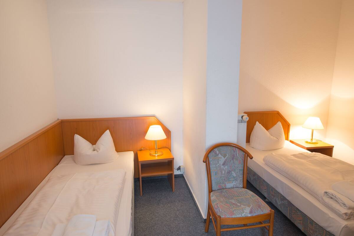 A hotel room with two single beds, a wooden nightstand with a lamp, a chair, and a small table.