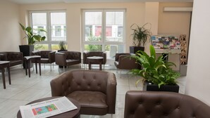 Um lobby moderno com poltronas de couro, uma mesa de centro com um jornal, janelas amplas e plantas em vasos.